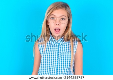 little kid girl with glasses wearing plaid shirt over blue background having stunned and shocked look, with mouth open and jaw dropped exclaiming: Wow, I can't believe this. Surprise and shock