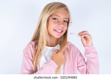 Little Kid Girl Wearing Pink Jacket Over White Background Holding An Invisible Aligner And Pointing At It. Dental Healthcare And Confidence Concept.