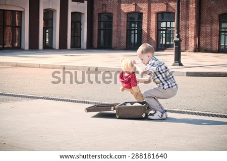 little kid is opening the suitcase on the sunny street in the morning, holding teddy bear