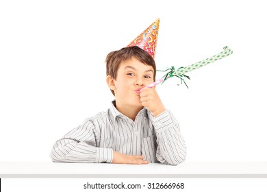 Little Kid With Party Hat Sitting At A Table And Blowing A Favor Horn Isolated On White Background