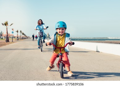 Little Kid Riding A Balance Bike With His Mother On A Bicycle In A City Park