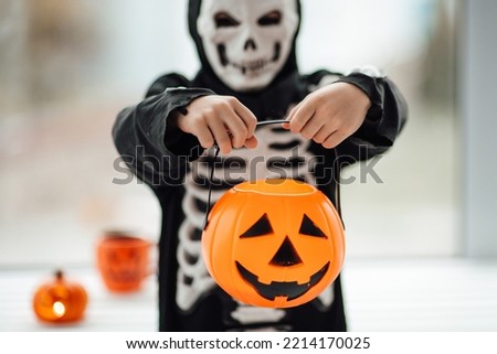 Little kid in skeleton costume with pumpkin candy bucket playing at home. Child is ready for trick or treat and scary gesture with cup of head Jack celebrates Halloween holiday party