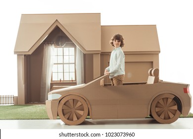 Little Kid Standing In Cardboard Car In Front Of Cardboard House Isolated On White