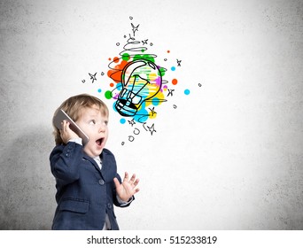 Little Kid Is Talking On The Phone Standing Near The Concrete Wall With Colorful Light Bulb Sketch On It. Concept Of A Bright Idea