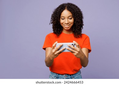 Little kid teen girl of African American ethnicity she wear orange t-shirt use play racing app on mobile cell phone isolated on plain pastel light purple background studio. Childhood lifestyle concept - Powered by Shutterstock