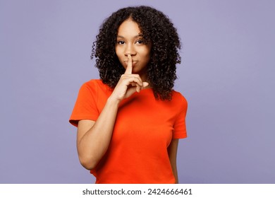 Little kid teen girl of African American ethnicity wear orange t-shirt say hush be quiet with finger on lips shhh gesture isolated on plain pastel light purple background. Childhood lifestyle concept - Powered by Shutterstock