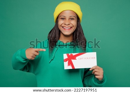 Little kid teen girl of African American ethnicity 13-14 years old wear hoody hat hold point on gift certificate coupon voucher card for store isolated on plain dark green background Childhood concept