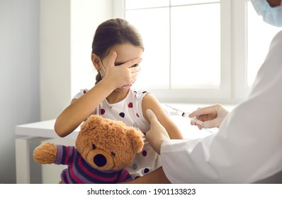 Little Kid Terrified By Injection At The Hospital. Girl Afraid Of Syringe Needle Covers Face While Getting Flu Vaccine At Pediatric Clinic. Doctor Giving A Shot To Scared Child Holding Teddy Bear Toy