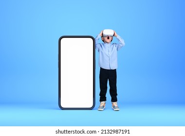 A little kid in virtual reality glasses, full length. Smartphone mock up copy space screen, blue background. Concept of metaverse and new device - Powered by Shutterstock