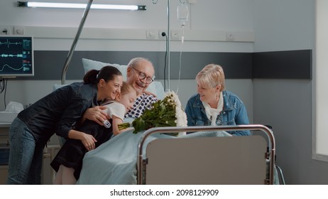 Little Kid And Woman Bringing Flowers To Old Patient At Family, Sitting In Hospital Ward Bed. Mother With Child Visiting Retired Man With Sickness In Intensive Care. Visitors Giving Support