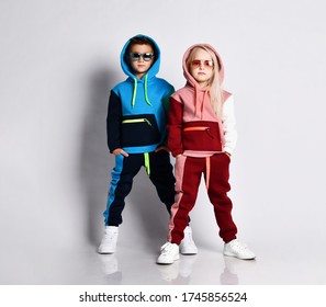 Little kids, boy and girl, in sunglasses and hoods, colorful tracksuits, sneakers. They posing isolated on white studio background. Childhood, fashion, advertising and sport.