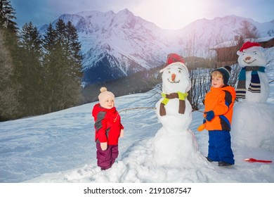 Little Kids Boy With Sister Girl Build, Dress Snowman Using Scarf And Hat Outside Over Mountain Peaks On Background