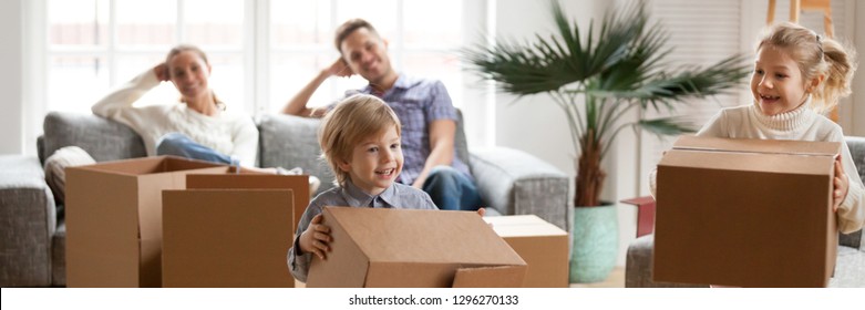 Little Kids Help With Stuff In Big Carton Boxes Play Having Fun, Happy Parents Resting On Couch In Living Room, Relocate Move Day At New Home Concept. Horizontal Photo Banner For Website Header Design