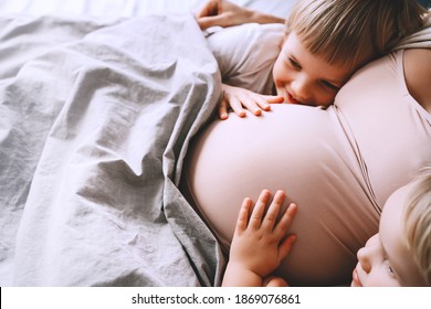Little kids hold hands on pregnant belly of mom. Pregnant woman with her children relaxing in bed. Loving mother and toddlers together at home. Third pregnancy. Maternity, family, parenting concept.
