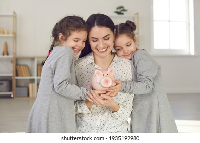 Little Kids Learning To Value Money. Happy Young Single Mother And Two Cute Daughters Holding Piggy Bank And Smiling. Saving Up For Children's Education, Family Finance, And Budget Management Concept