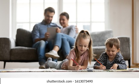 Little Kids Lying On Floor In Living Room Paint Picture With Colorful Pencils, Parents Relax On Couch Enjoy Weekend Together, Small Children Brother And Sister Drawing At Home, Family Time Concept