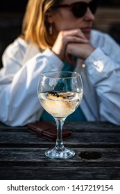 Little Kingshill, Buckinghamshire/UK - May 25 2019: Young Woman Sits Behind A Glass Of Gin And Tonic. She Is Wearing A White Jacket And Sunglasses. Hands Are Clenched In Front Of Her Face. Outside 