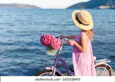 Little Lady With Bicycle And Pink Flowers On Sea Beach. Kid Girl In Straw Hat And Dress Is Riding On Coastline. Hydrangea Is In Bike Basket. Concept Of Tenderness, Childhood, Romantic Summer.