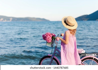Little Lady With Bicycle And Pink Flowers On Sea Beach. Kid Girl In Straw Hat And Dress Is Riding On Coastline. Hydrangea Is In Bike Basket. Concept Of Tenderness, Childhood, Romantic Summer Holidays.
