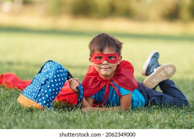 little Latin American schoolboy in superhero costume with red carpet is lying with satchel on lawn in fresh air. child fantasizes and pretends to be a superhero. A place for text or advertising.