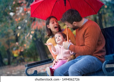 Little Lover Of Rain Smiling With Her Happy Family Under Autumn Rain. Smiling Parents And Little Child Girl Have Fun Time While Playing Outdoor Under Autumn Shower. Happy Family Under Umbrella.
