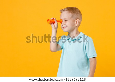 Little male kid boy 5-6 years old wearing stylish blue turquoise t-shirt polo looking far away distance through toy spyglass telescope isolated on yellow color wall background, child studio portrait