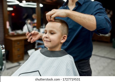 Little Man. Cute Boy Sitting In A Barbershop Chair And Smiling While Male Barber Styling His Hair. Barbershop. Child Visiting Barber Salon. Kids Haircut