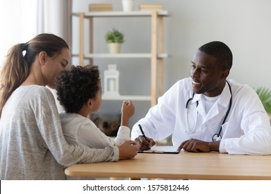 Little mixed race cute boy sitting on mommys knees talking to smiling african american male doctor at clinic. Cheerful kind pediatrician carefully listening to small kid patient, writing down notes.