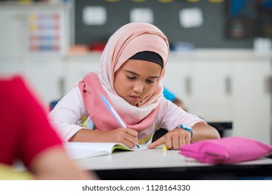 Little muslim girl wearing hijab and doing homework in class. Young arab schoolgirl in chador writing during exam in classroom. Pretty elementary child writing notes during lesson at school.