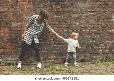 Little nephew pulling her elder aunt to go further. Young girl in checked shirt, black trousers standing at place near brick wall. Tiny kid in jeans and stripped sweater wants her aunt to follow him. - Shutterstock ID 495872131