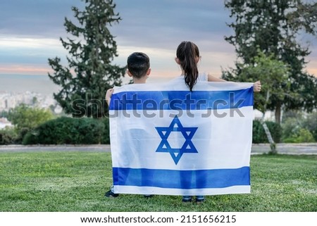 Little patriot Jewish children standing with the flag of Israel enjoying view of city. Back view image of young kids covering themselves with flag of Israel on meadow at sunset.