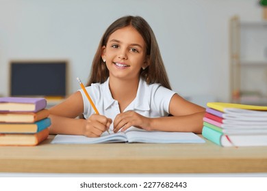 Little positive girl teenager elementary school student posing at desk with textbooks and notebooks holding pencil in hand and looking at camera, sits in classroom. Back to school, primary education - Powered by Shutterstock