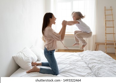 Little preschool daughter frolics jumping on bed holding cheerful mothers hands, after daytime or night sleep girl and her mom spend active carefree time do exercise or just fool around in the bedroom