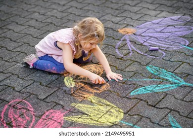 Little preschool girl painting with colorful chalks flowers on ground on backyard. Positive happy toddler child drawing and creating pictures on asphalt. Creative outdoors children activity in summer.