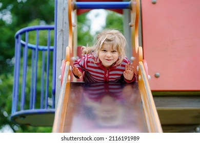 Little Preschool Girl Playing On Outdoor Playground. Happy Toddler Child Climbing And Having Fun With Summer Outdoors Activity. Girl Slinding Down The Slide.