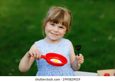 Little Preschool Girl Playing With Toy Kitchen In Garden. Happy Toddler Child Having Fun With Role Activity Game Preparing Food. Children Play Outdoors In Summer