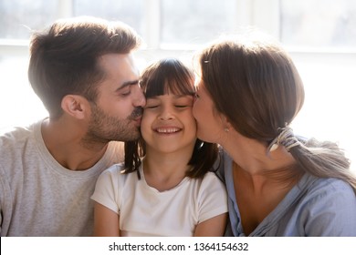 Little preschool girl sitting with mother and father closed eyes smile feels happy while her parents kissing her on cheek expressing love care support and devotion, well being friendly family concept