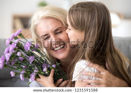 Little preschool granddaughter kissing happy older grandma on cheek giving violet flowers bouquet congratulating smiling senior grandmother with birthday, celebrating mothers day or 8 march concept
