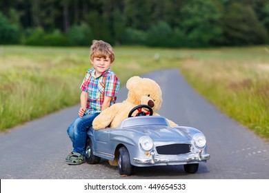 Little Preschool Kid Boy Driving Big Toy Car And Having Fun With His Plush Toy Bear, Outdoors. Child Enjoying Warm Summer Day In Nature Landscape