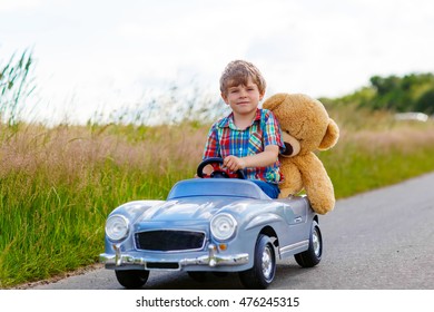 Little Preschool Kid Boy Driving Big Toy Car And Having Fun With Playing With His Plush Toy Bear, Outdoors. Child Enjoying Warm Summer Day In Nature Landscape