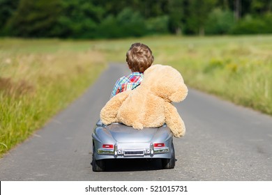 Little Preschool Kid Boy Driving Big Toy Car And Having Fun With Playing With His Plush Toy Bear, Outdoors. Child Enjoying Warm Summer Day In Nature Landscape