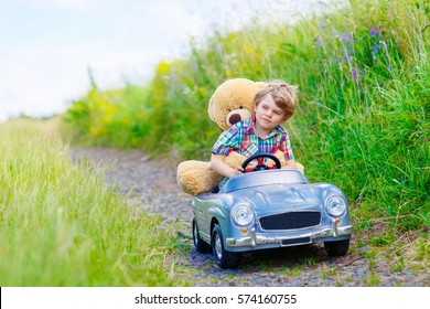 Little Preschool Kid Boy Driving Big Toy Car And Having Fun With Playing With His Plush Toy Bear, Outdoors. Child Enjoying Warm Summer Day In Nature Landscape