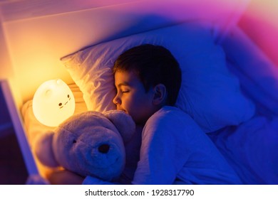 Little preschool kid boy sleeping in bed with colorful night led lamp. School child dreaming and holding plush toy. Kid angry of darkness.