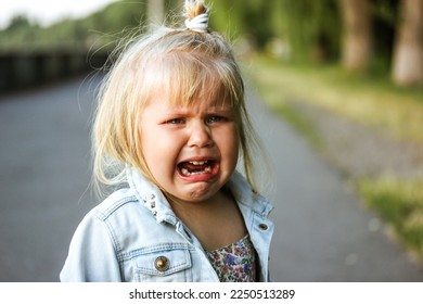 A little preschool-aged 3-5 year old girl is desperately crying loudly standing on a street. The child is lost. Negative emotions. White child refugee from Ukraine. Unhappy toddler kid from orphanage.