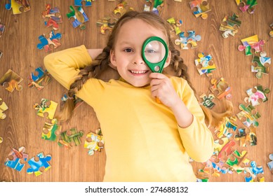Little pretty smiling girl in yellow pullover laying on the floor and playing with puzzle and loupe.