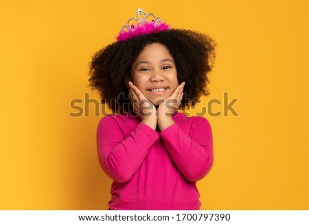 Little Princess. Portrait Of Cute Black Girl Posing To Camera With Crown On Head And Touching Her Face, Yellow Studio Background With Free Space