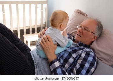 Little Red-haired Baby Sitting On Grandpa Belly. Senior Man Laying On Bed And Talking With Cute Toddler. Help From Grandparents And Staying At Home Concept