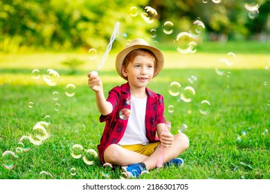 a little red-haired boy inflates soap bubbles on a green lawn in a hat and a red shirt in summer having fun playing - Powered by Shutterstock