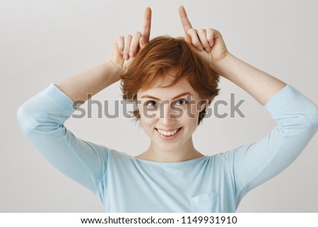 Little redhead devil makes guy fall for her from first sight. Portrait of attractive european woman with ginger hair making horns on top of head with index fingers and smiling with interest.