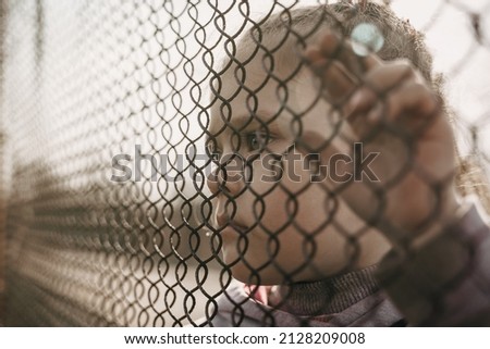 A little refugee girl with a sad look behind a metal fence. The social problem of refugees and internally displaced persons.